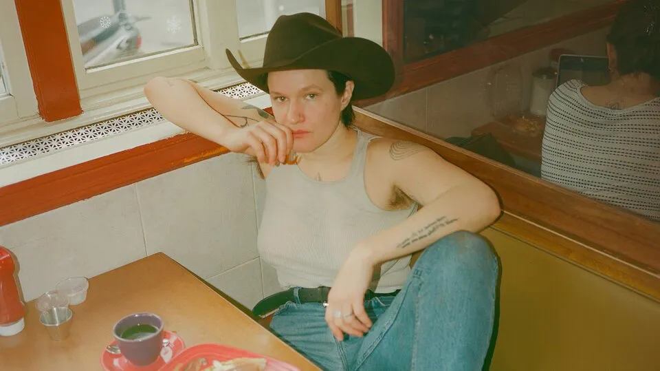 Adrianne Lenker, a white woman wearing a cowboy hat and a tank top, leaning against a diner booth and looking at the camera.