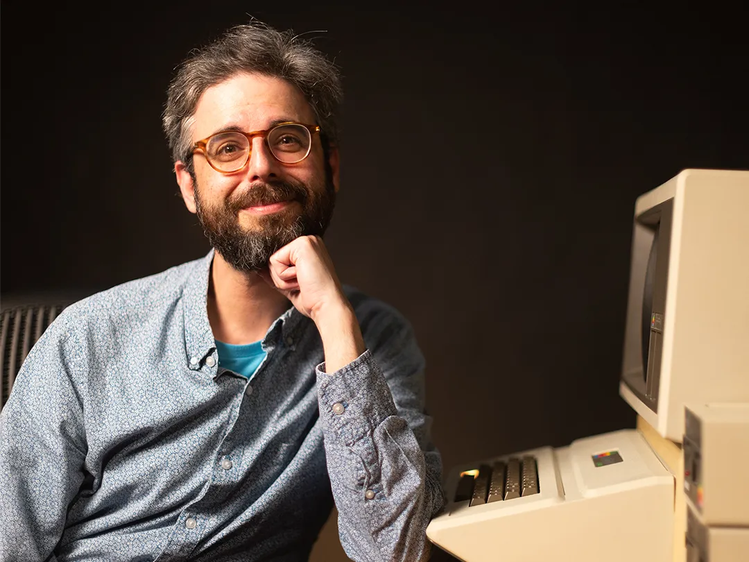 A photo Daniel Shiffman, creator of The Coding Train. He is a middle-aged white man with a dark, full beard and gentle smile. He is leaning against an old macintosh computer, looking at the photographer.